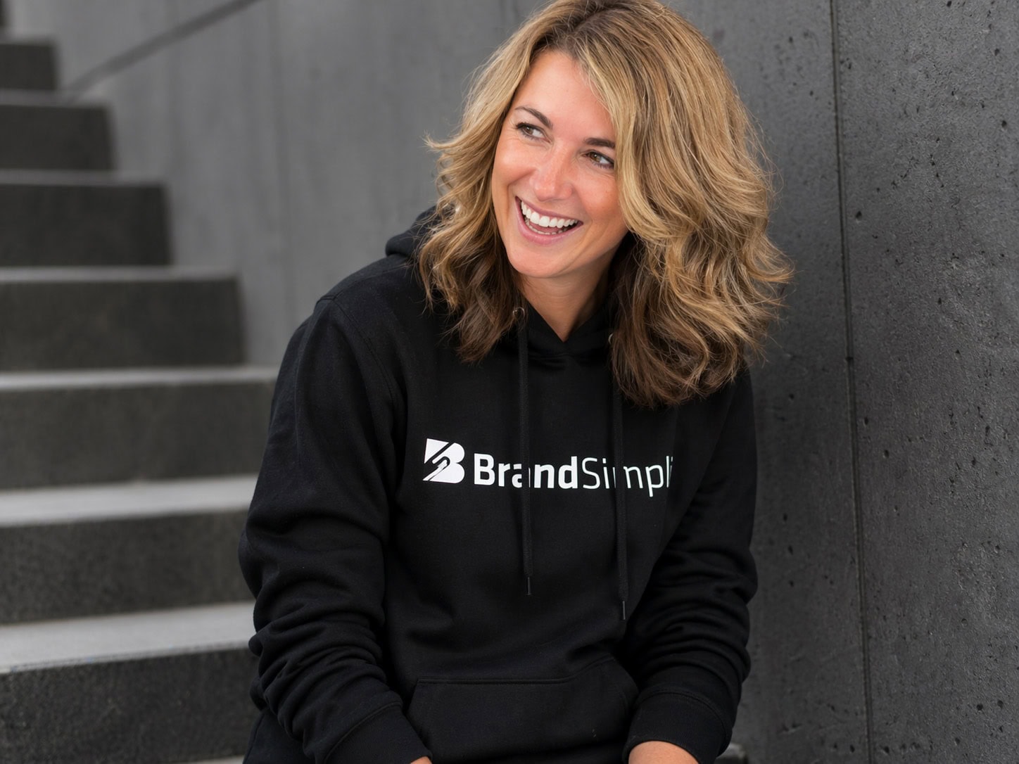 Frau trägt schwarze Hoodie mit BrandSimpli Logo, lächelnd, urbaner Hintergrund.