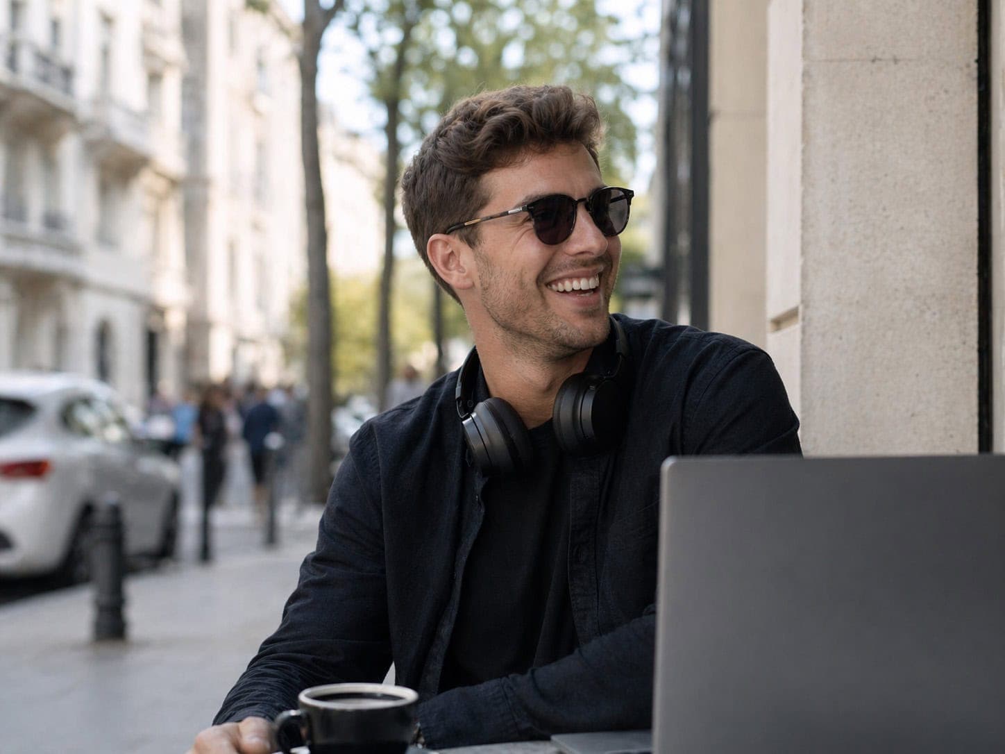 Junger Mann mit Laptop, Sonnenbrille und Kopfhörern, arbeitet draußen in der Stadt.