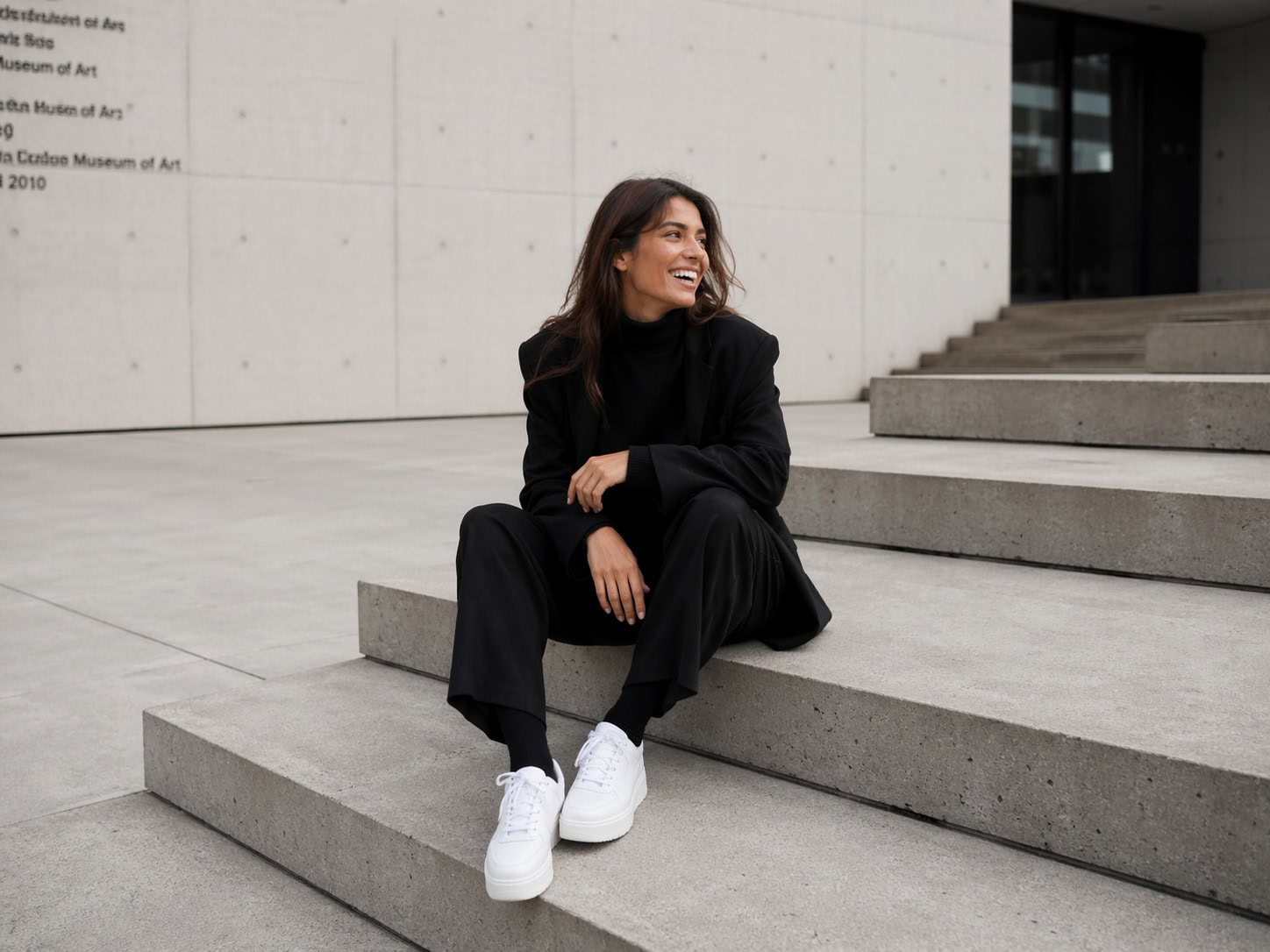 Junge Frau sitzt auf Treppe vor moderner Architektur, lächelnd, casual Style.
